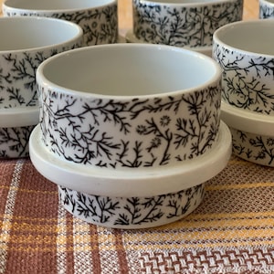 Puede incluir: Juego de seis tazas de cerámica blanca con un patrón floral negro. Las tazas están apiladas una encima de la otra.