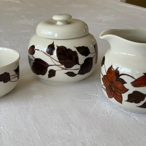 May include: A set of three white ceramic sugar and creamer containers with brown leaf and flower designs.