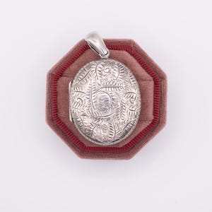 Peut inclure: Un médaillon ovale en argent avec un motif floral complexe, présenté sur un coussin de velours rouge.