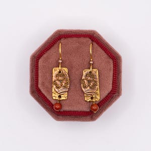 May include: A pair of gold-toned earrings with rectangular pendants featuring an ornate, swirling design. Each earring has a small, round, reddish-brown bead at the bottom and is displayed on a maroon velvet jewelry box.