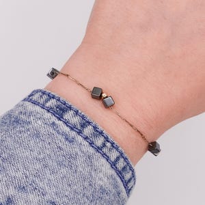 Puede incluir: Una pulsera delicada con pequeñas cuentas cuadradas de color gris oscuro y una sola cuenta dorada ensartadas en una fina cadena dorada. La pulsera se muestra en una muñeca, con una manga de camisa vaquera en el fondo.