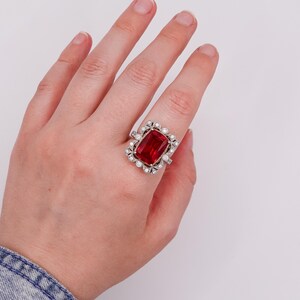 May include: A silver-toned ring featuring a large, rectangular, faceted red gemstone at the center. The stone is surrounded by a decorative border of small, round, clear stones set in a metallic frame that extends across the finger.