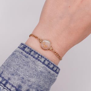 Puede incluir: Una pulsera de cadena en tono dorado con un colgante circular que tiene un centro translúcido de color rosa pálido con pequeñas escamas doradas dispersas. La pulsera se asegura con una delicada cadena de eslabones metálicos alrededor.