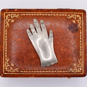 May include: A silver-toned metal hand-shaped object featuring engraved lines on the fingers and a heart-shaped design on the palm, resting on a rectangular brown leather box with gold-colored floral border detailing along the edges.