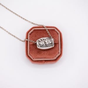 May include: A silver-toned necklace with a rectangular pendant featuring a detailed relief design. The pendant rests on a pink velvet jewelry box. The chain is delicate and the overall aesthetic is vintage.