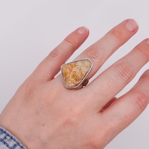 Könnte beinhalten: Ein Ring mit einem großen, unregelmäßig dreieckigen fossilen Korallenstein, der ein beigefarbenes und cremefarbenes Sternmuster aufweist und in eine glatte, polierte silberfarbene Metallschiene gefasst ist, die den Finger umschließt.