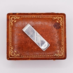 Könnte beinhalten: Ein silberner Geldklammer mit geometrischem Design liegt auf einer braunen Lederbox mit goldenem Blumenschmuck. Die rechteckige Box hat ein leicht abgenutztes Aussehen.