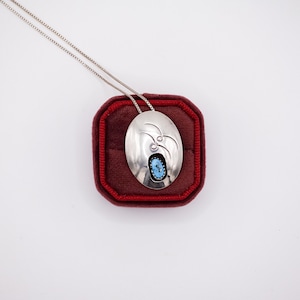 May include: A silver pendant necklace with an oval pendant featuring a turquoise stone and etched bird designs. The pendant is displayed on a red velvet jewelry box. The chain is a delicate silver chain.