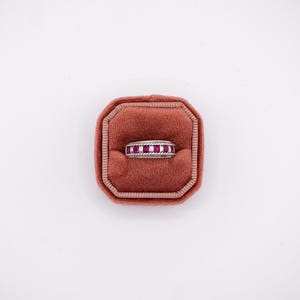 May include: A silver band ring featuring a row of square-cut, deep pinkish-red rubies set between two twisted rope-style metal borders. The ring is displayed centered within a square, rust-colored velvet ring box with rounded corners.