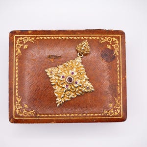 Puede incluir: Un colgante dorado con un diseño floral y pequeñas piedras preciosas rojas descansa sobre una caja de cuero marrón. La caja tiene un borde floral dorado. El colgante mide aproximadamente 5 cm de largo.