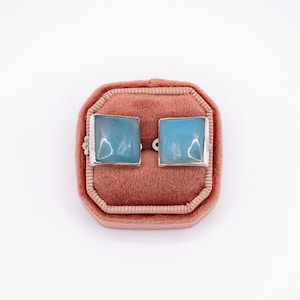 May include: A pair of square-shaped cufflinks with a light blue gemstone set in a silver metal frame. The cufflinks are displayed inside a pink velvet jewelry box against a white background. The gemstones have a translucent appearance.