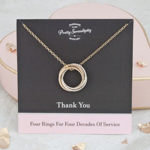 May include: Gold necklace with four interlocking rings, each ring a different colour. The necklace is displayed on a black card with the text "Thank You" and "Four Rings For Four Decades Of Service".
