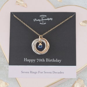 May include: A gold necklace with a pendant of seven interlocking rings and a blue gemstone. The necklace is displayed on a black card with the text "Happy 70th Birthday" and "Seven Rings For Seven Decades".