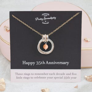 May include: A gold necklace with a silver pendant featuring three larger rings and five smaller rings, a coral-colored bead, and the text "Happy 35th Anniversary". The card reads "Three rings to remember each decade and five little rings to celebrate your special 35th year."
