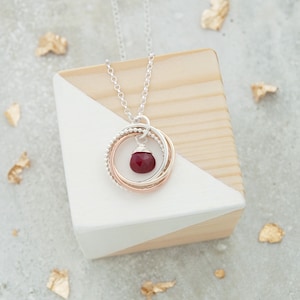 May include: A silver chain necklace with a pendant featuring three interlocking circles in silver, rose gold, and gold, with a red gemstone in the center.