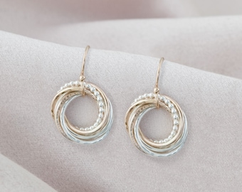 May include: A pair of gold and silver earrings with a circular design. The earrings are made of multiple intertwined rings, creating a textured and intricate look.
