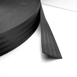 Könnte beinhalten: Schwarzes Gurtband mit drei schwarzen Streifen, die in Längsrichtung verlaufen. Das Band ist aufgerollt und ein Abschnitt ist ausgezogen, um die Textur zu zeigen.