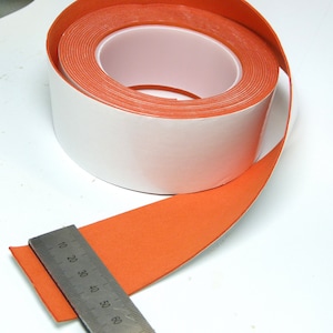 May include: A roll of orange double-sided tape with a white backing. The tape is 2.5 cm wide and is measured with a ruler.