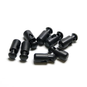 May include: A group of eight black plastic cord locks, also known as toggle stops, are scattered on a white surface. These cord locks are used to secure cords and ropes.