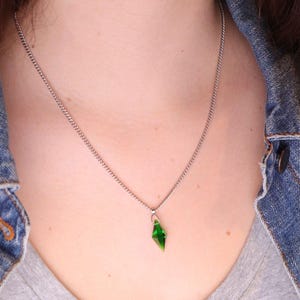 Pode incluir: Um colar de corrente prateada com um pequeno pendente verde esmeralda em forma de diamante. O pendente é facetado, captando a luz. O colar é usado sobre uma camiseta cinza e uma jaqueta jeans.