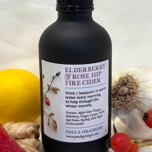 May include: A black glass bottle with a black label that reads "Elderberry & Rose Hip Fire Cider". The label also includes instructions for use and a list of ingredients. The bottle is sitting on a white surface with a red chili pepper, a lemon, and a ginger root.