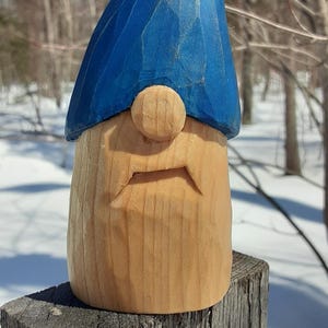 May include: A hand-carved wooden gnome sculpture with a blue hat and a light brown face. The gnome has a round nose and a carved mouth. The sculpture is displayed outdoors on a wooden post, with a snowy background.