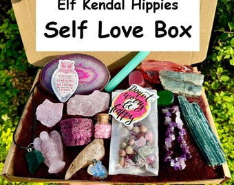 May include: A pink and green self-love box with crystals, a pink angel figurine, a green stone necklace, a small bottle of pink crystals, a donut sticker that says "Don't worry be happy", and a sticker with an owl and the text "Knowledge comes from learning, wisdom comes from living".