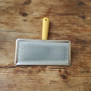 May include: A silver grooming brush with a yellow plastic handle. The brush has a rectangular shape and is filled with rows of metal bristles.