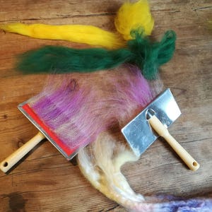 May include: Two felting tools with wooden handles and metal surfaces. A variety of colorful wool fibers are spread out on a wooden surface.