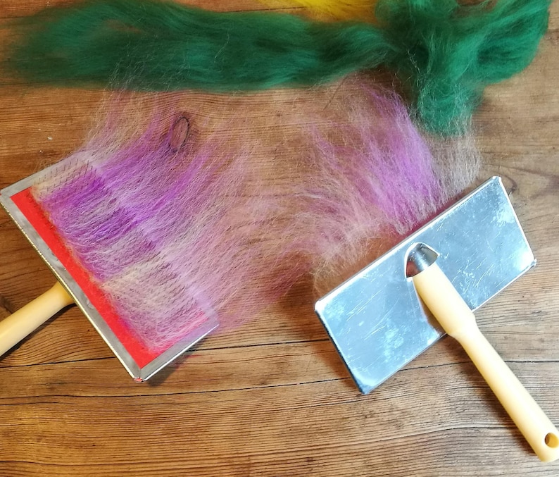 May include: Two felting tools with wooden handles. One tool has a red base and the other has a silver base. A strand of pink and purple wool is being felted on the red tool.