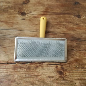 May include: A silver grooming brush with a yellow plastic handle. The brush has a rectangular shape and is filled with rows of metal bristles.