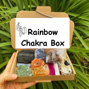 May include: A cardboard box labeled "Rainbow Chakra Box" containing various colorful crystals and stones. The box is open, revealing a collection of natural gemstones in shades of black, blue, orange, green, and pink. A small illustration of a cartoon elf is on the box.