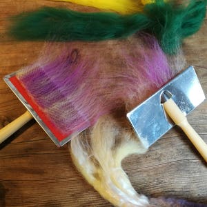 May include: Two wooden handled tools for felting wool. One tool has a red felt surface and the other has a metal surface. There are four different colors of wool on the wooden surface.