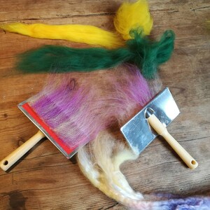 May include: Two felting tools with wooden handles and metal surfaces. A variety of colorful wool fibers are spread out on a wooden surface.