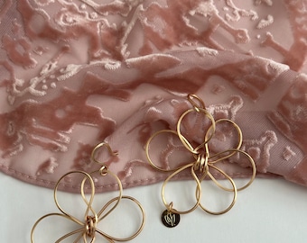 Puede incluir: Pendientes de oro en forma de flor con un delicado diseño de alambre. Los pendientes cuelgan de un fondo de terciopelo rosa.