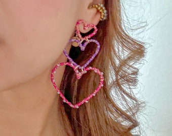 Puede incluir: Un par de pendientes colgantes con tres corazones superpuestos. Los corazones están hechos de hilo rosa y morado envuelto alrededor de un marco de alambre.
