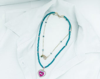 Puede incluir: Dos collares en capas con cuentas turquesas y detalles dorados. El collar superior tiene pequeños amuletos de ojo turco. El collar inferior presenta un colgante en forma de corazón con un diseño de ojo en rosa y blanco. Los collares se muestran sobre una camisa blanca.