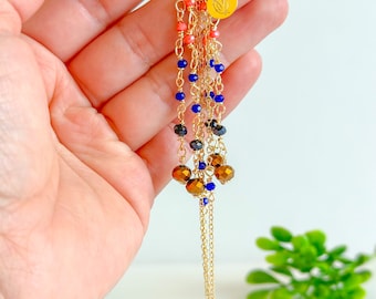 Puede incluir: Pendientes de aro dorados con un diseño multi-cadena que presenta cuentas de coral, azul y marrón. Los pendientes tienen un pequeño dije dorado con un diseño floral.