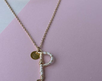 Puede incluir: Un collar de cadena dorada con un hilo blanco envuelto alrededor de un colgante de la letra 'P' dorada. Un pequeño disco dorado con una letra 'V' grabada en él cuelga sobre la 'P'.