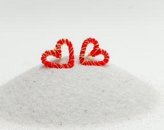 Puede incluir: Dos pendientes de alambre en forma de corazón rojo con detalles dorados sobre una superficie de arena blanca.
