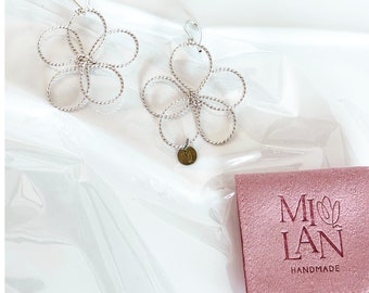 Puede incluir: Un par de pendientes plateados con forma de flor y diseño de alambre retorcido. Cada pendiente tiene un pequeño dije redondo. También se ve una bolsa rosa con el texto "MI LAN HANDMADE".