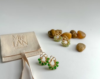 Puede incluir: Un par de pendientes de alambre dorado con cuentas de vidrio verde. Los pendientes se muestran sobre una superficie blanca con pequeñas piedras.