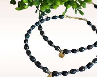 Puede incluir: Dos delicados collares con cuentas de perlas azul oscuro y dijes dorados. Los collares están superpuestos y presentan un diseño sencillo.