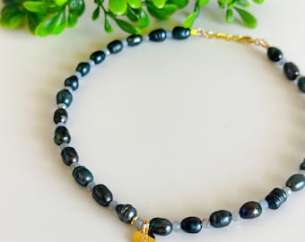 Puede incluir: Un delicado collar gargantilla hecho con perlas de agua dulce azul oscuro y un pequeño dije de oro. El collar está sobre una superficie blanca con follaje verde en el fondo.