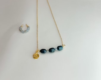 Puede incluir: Un collar de cadena dorada con un pequeño dije dorado y una fila de cuentas azul oscuro. Un par de aretes de aro pequeños a juego con cuentas azul claro.