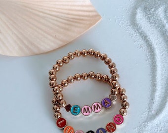 Puede incluir: Dos pulseras de cuentas doradas con cuentas de letras de colores que deletrean los nombres "Emma" y "Luciano".
