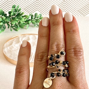 May include: A gold ring with black beads and a charm. The ring is worn on a finger with a light pink nail polish.