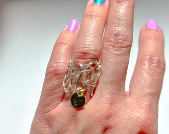 Puede incluir: Un anillo de alambre plateado con un dije de oro colgando. El anillo se usa en un dedo.