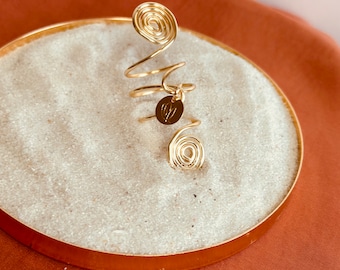 Puede incluir: Anillo de espiral dorado con un pequeño dije redondo. El anillo está hecho de alambre y tiene un diseño único.