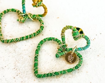 Puede incluir: Un par de pendientes en forma de corazón verde y dorado con un pequeño dije dorado que dice "SL".